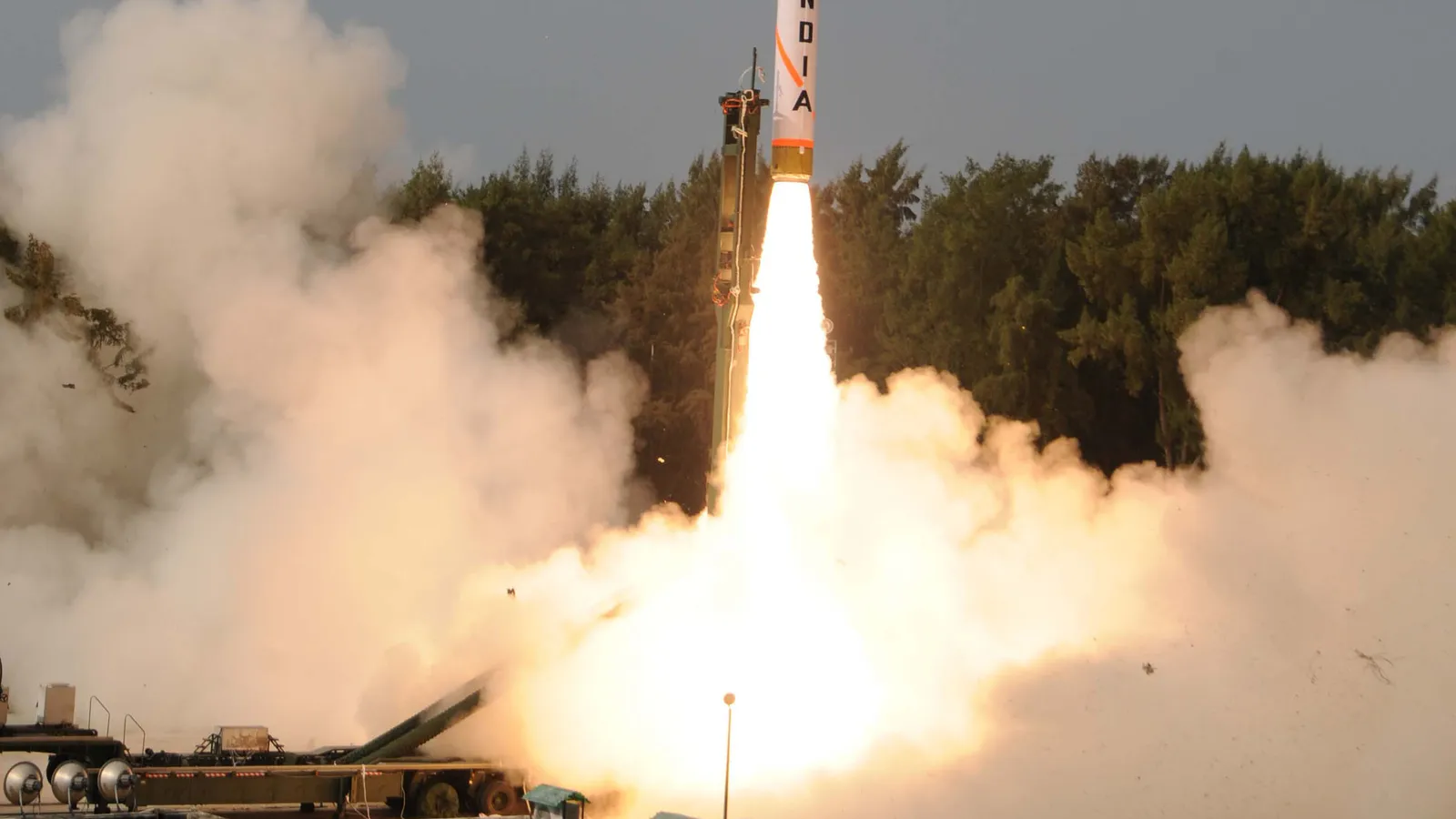 An Agni-IV ballistic missile lifting off from its road-mobile launcher in a cloud of exhaust during a test flight on the Odisha coast, with the tricolour-liveried missile body rising past a treeline.