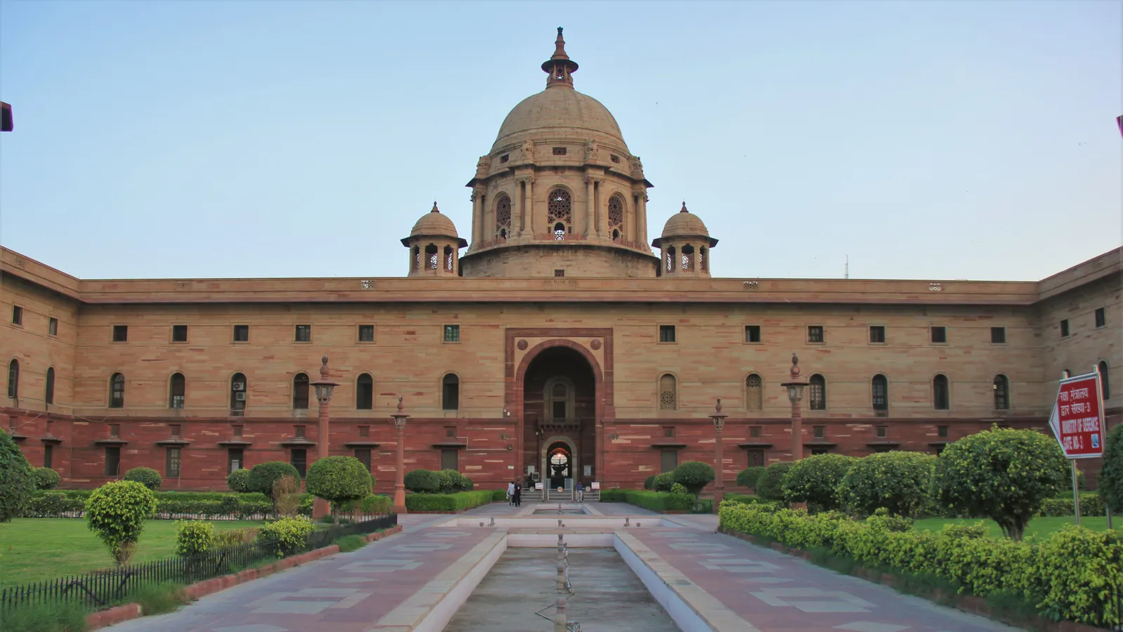 The sandstone facade and central dome of South Block on Raisina Hill, New Delhi, which houses the Ministry of Defence and Army Headquarters, seen from its front lawns with a Ministry of Defence gate sign at right.