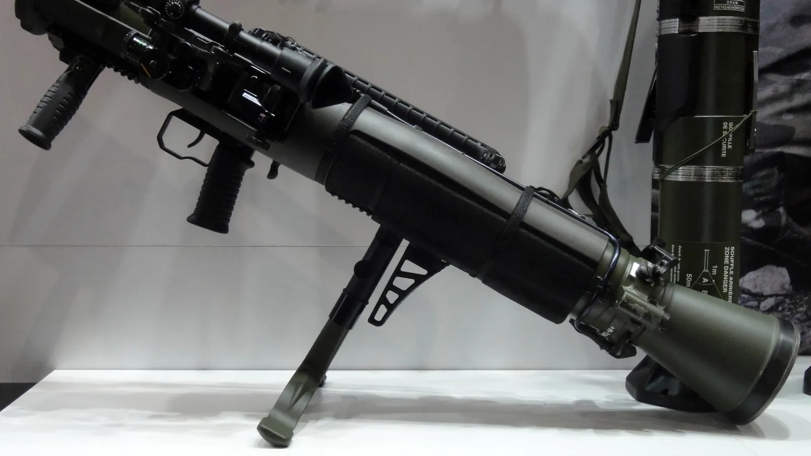 A Carl Gustaf M4 84mm recoilless launcher on display, resting on its foregrip stand with an olive-green 84mm ammunition tube standing beside it.