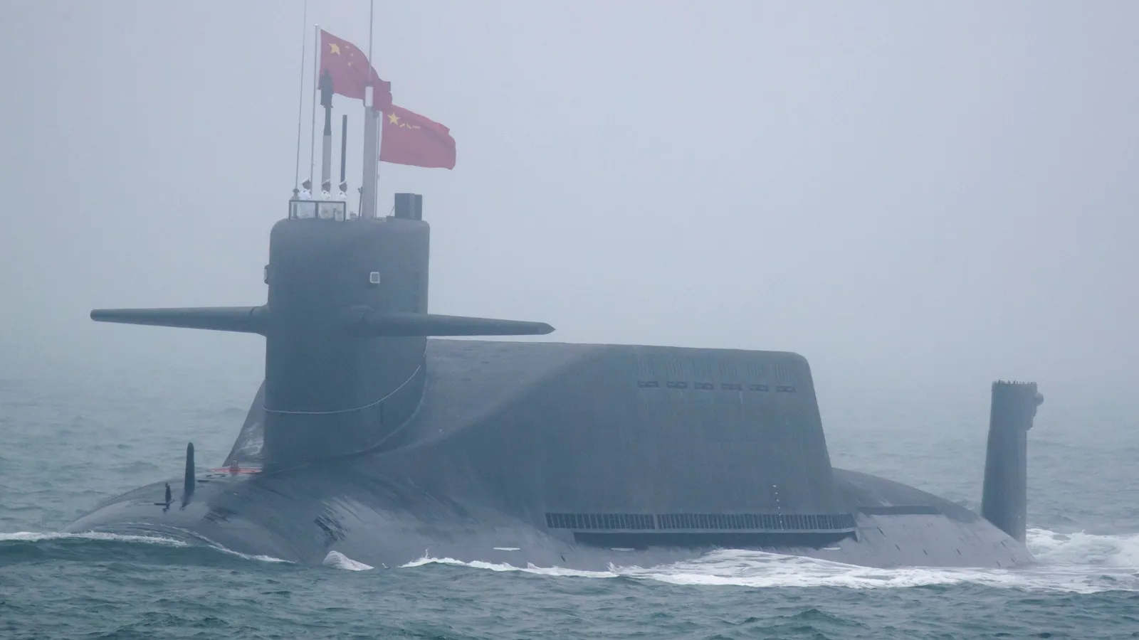 A Chinese Type 094 Jin-class nuclear-powered ballistic missile submarine sails on the surface in misty conditions, red PLA Navy flags flying from its sail and crew standing atop the conning tower.