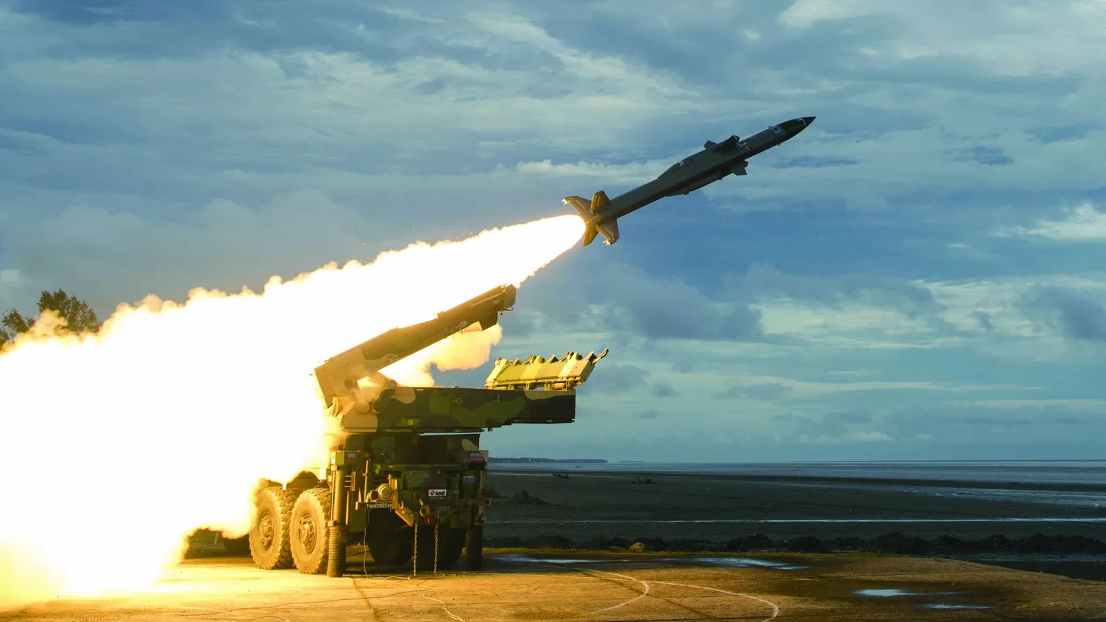 An Akash Prime surface-to-air missile lifts off in a plume of flame from a camouflaged truck-mounted launcher at a coastal test range.