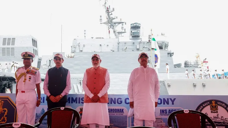Prime Minister Narendra Modi stands with a naval officer and dignitaries on a dais marked Tri Commissioning Ceremony, with a grey Indian Navy warship manned by sailors behind them.