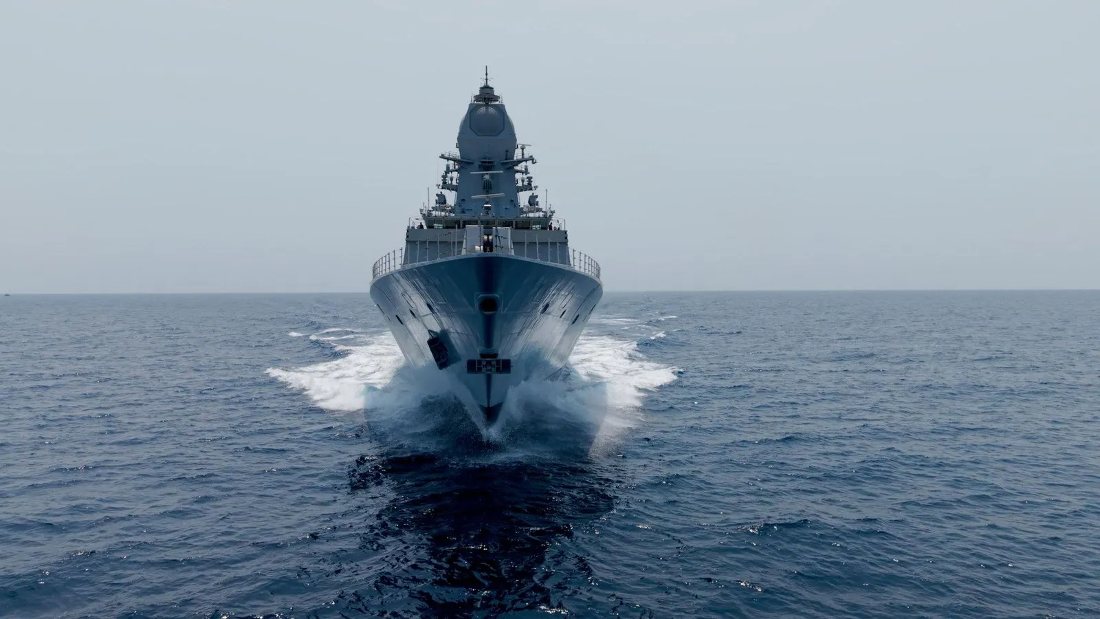 Bow-on view of the Project 17A stealth frigate INS Mahendragiri underway at sea, spray breaking around the hull as it cuts through calm water.