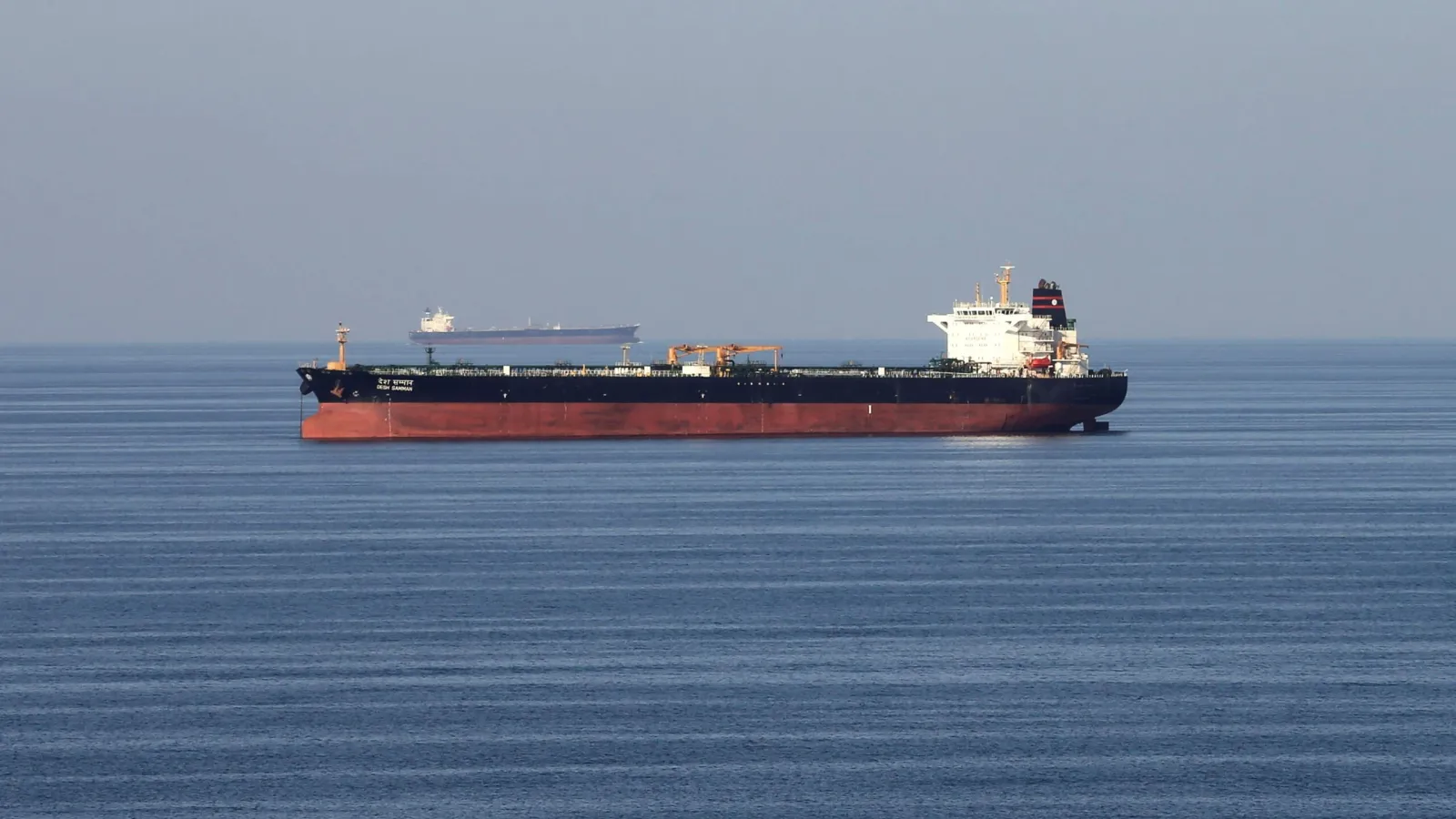 A laden crude oil tanker sails through calm waters of the Strait of Hormuz with a second tanker visible on the horizon.