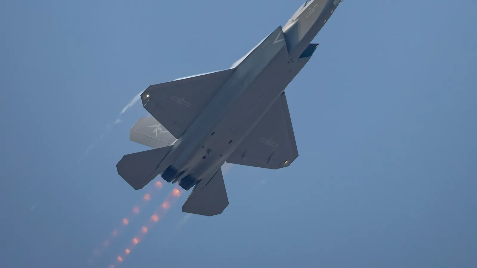 A grey Chinese J-35A stealth fighter climbs steeply with both afterburners lit, seen from below against a clear blue sky.