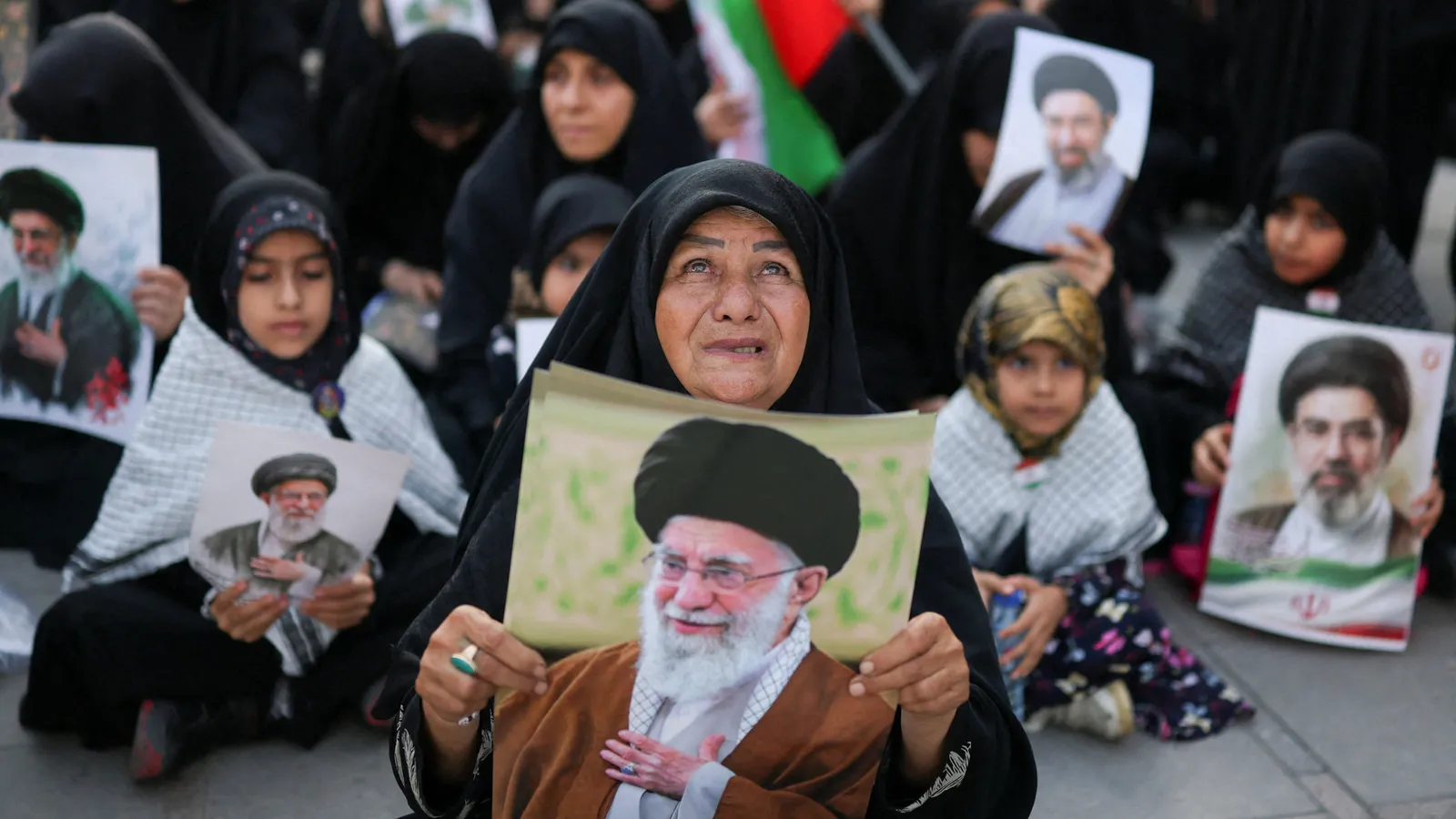 A weeping woman in a black chador holds a portrait of Ayatollah Ali Khamenei, surrounded by seated mourners holding similar posters and an Iranian flag, during funeral ceremonies in Tehran.