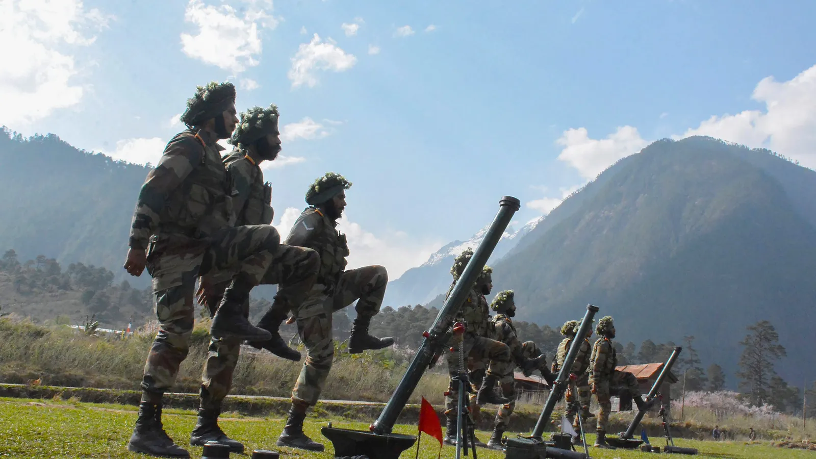 Indian Army soldiers with camouflaged helmets drill beside a row of mortars on a grass field in a Himalayan valley in Arunachal Pradesh.