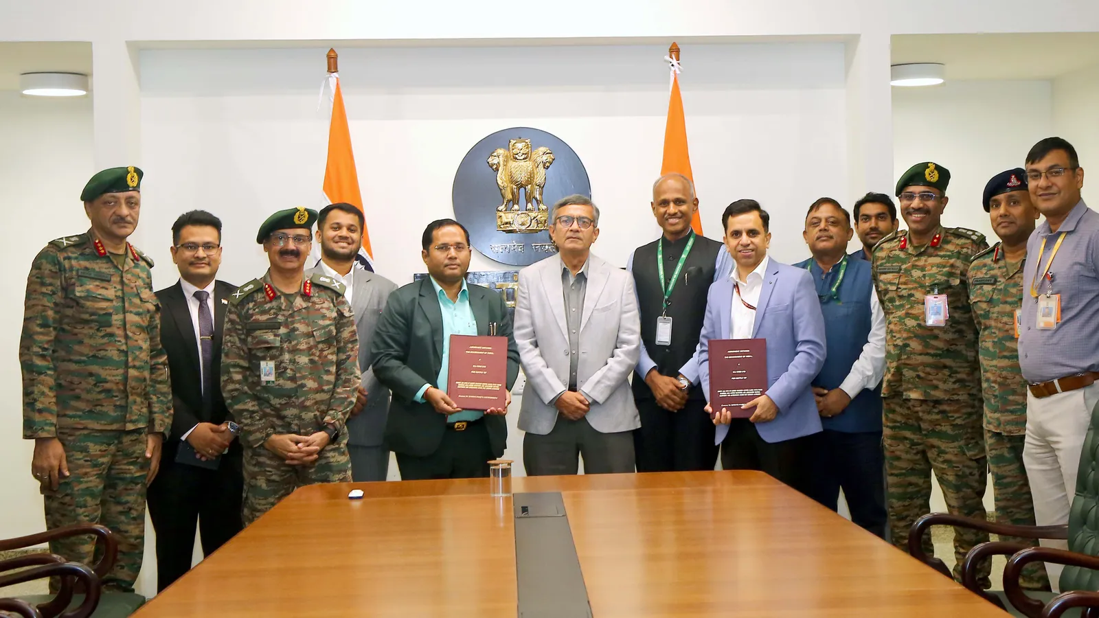 Defence Secretary Rajesh Kumar Singh stands at the centre of a row of Indian Army officers in combat uniform and civilian company representatives in a Ministry of Defence conference room, two representatives holding signed maroon contract folders, with the Ashoka emblem and two Indian flags on the wall behind them and a long wooden table in the foreground.