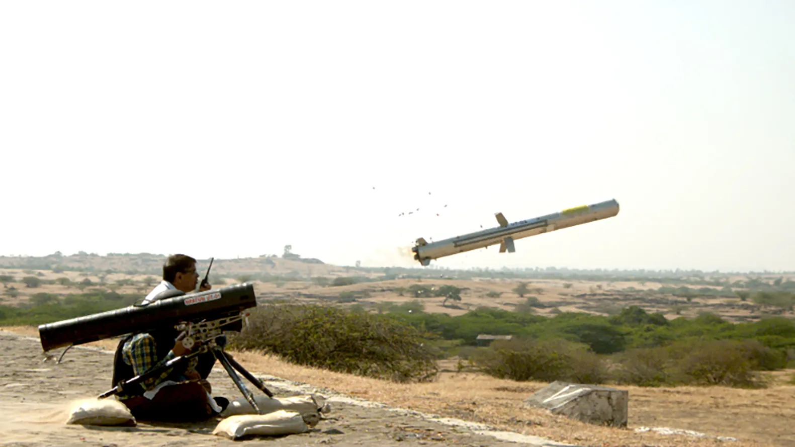 An MP-ATGM missile in mid-air moments after launch from a tripod-mounted tube, fired by a seated operator at a sandbagged position overlooking scrubland.