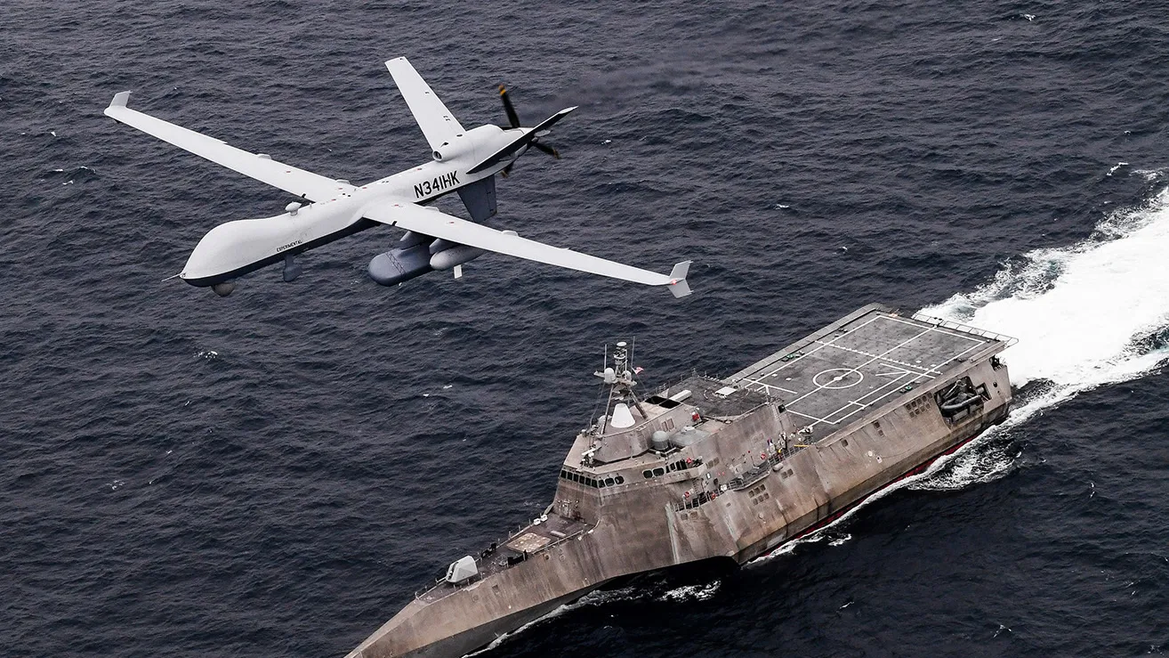 A General Atomics MQ-9 unmanned aircraft in maritime surveillance configuration flying low over an Independence-class littoral combat ship at sea, its long straight wings and rear propeller clearly visible against dark blue water.