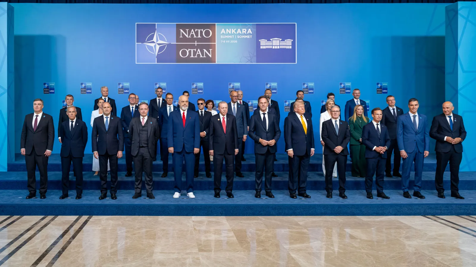 Heads of state and government of NATO's 32 member countries stand on a tiered platform for the official family photo at the Ankara summit, in front of a blue backdrop bearing the NATO Ankara Summit emblem.