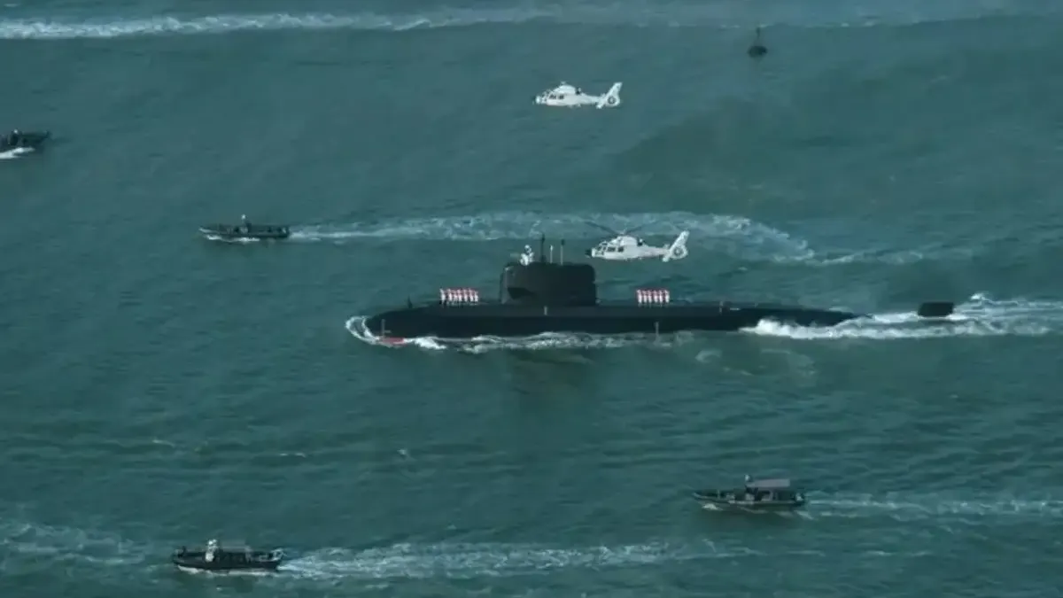 A Hangor-class submarine sails on the surface with crew lined up on deck, escorted by helicopters and small boats during its arrival at Karachi.