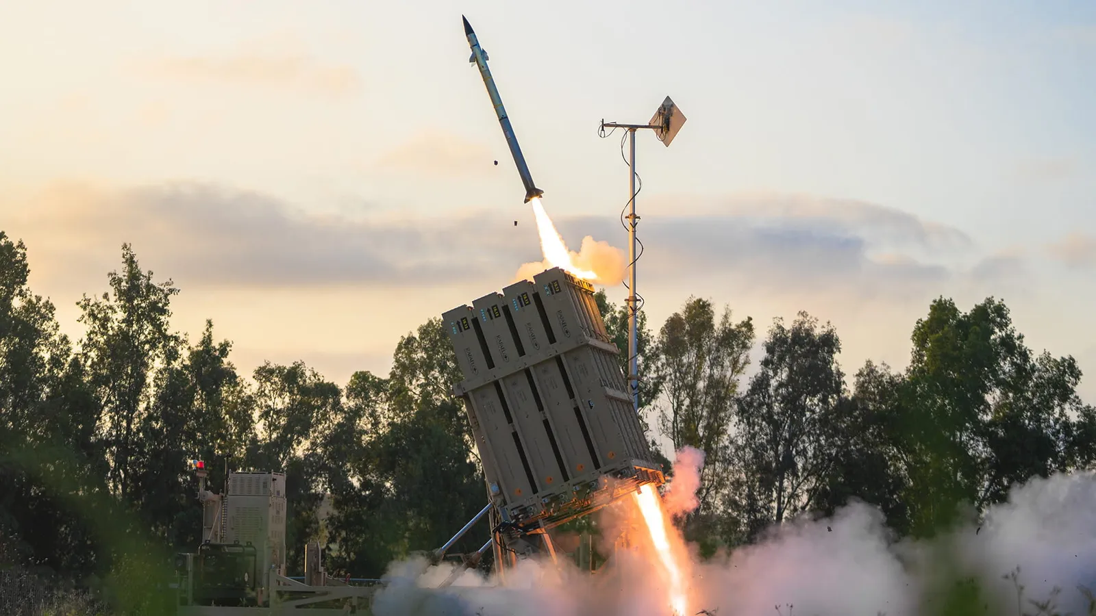 A Tamir interceptor climbing away from an angled Iron Dome launcher just after firing, with exhaust flame and smoke trailing below it against an evening sky.