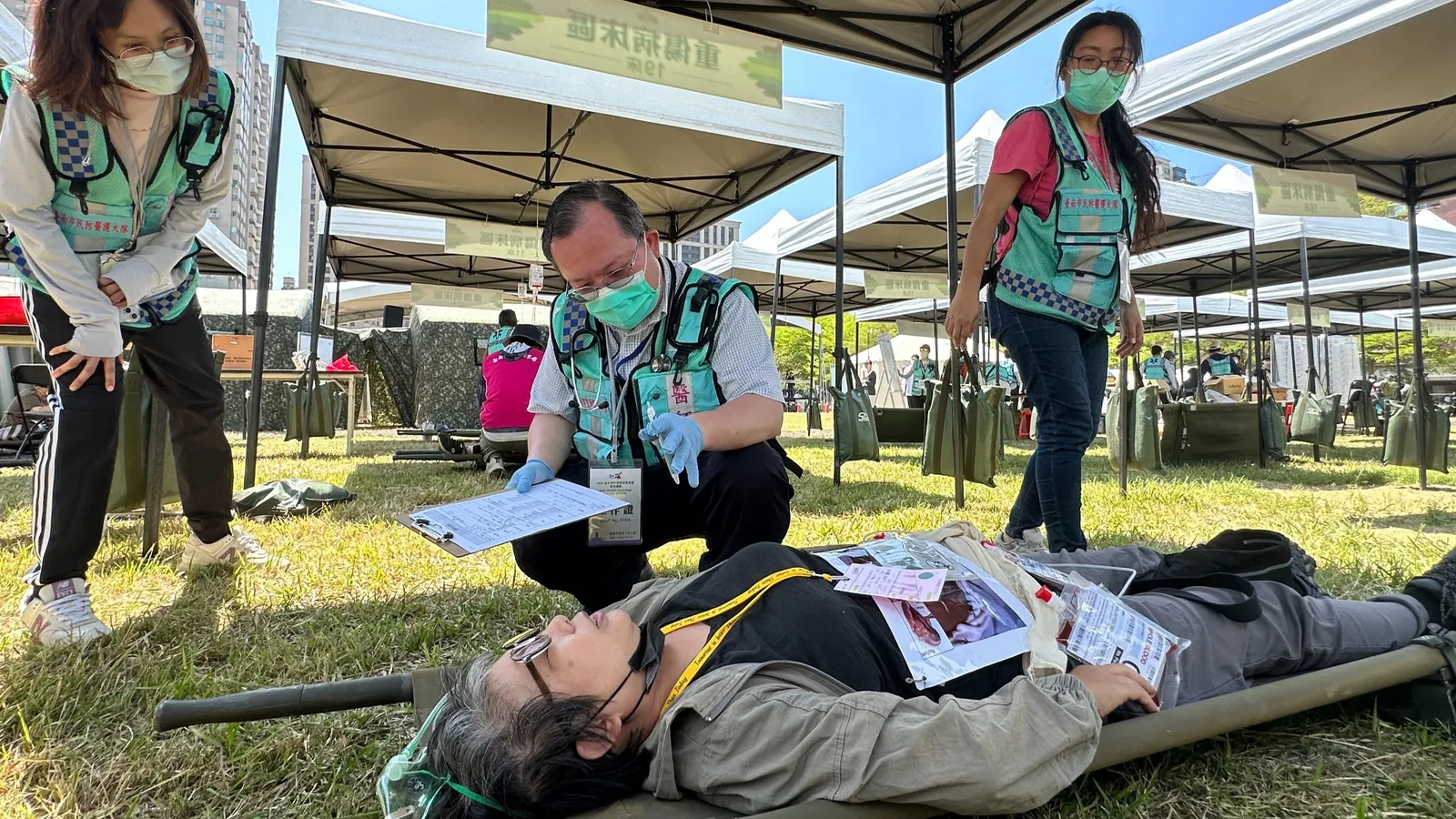 Civil-defence medical volunteers in green vests triage a volunteer playing a casualty on a stretcher under field tents during a Taiwanese civil-defence drill.