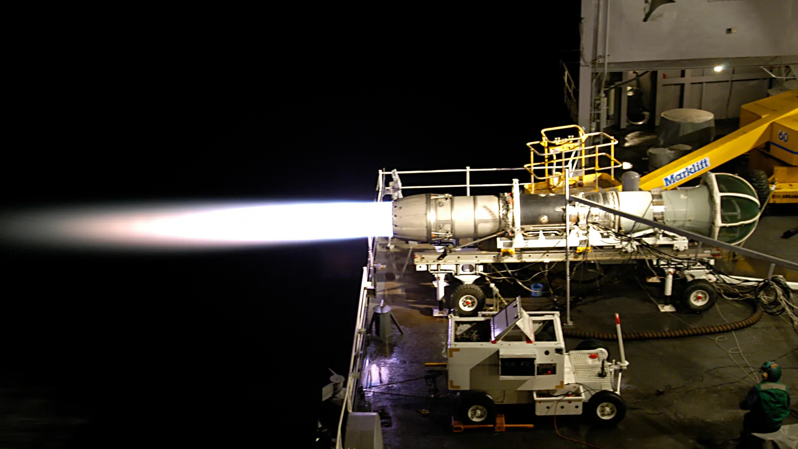 A General Electric F404 turbofan engine mounted on a wheeled test stand fires its afterburner during a night test, sending a bright exhaust plume off the deck.