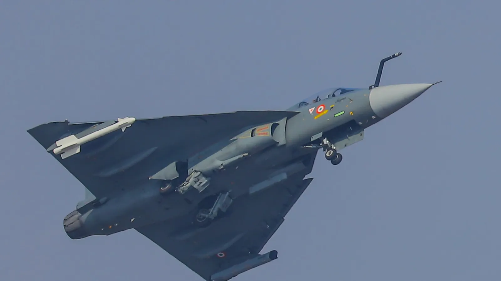 An Indian Air Force Tejas Mk1A light combat aircraft banking against a clear sky with landing gear extended and a wingtip missile fitted, during its flying display at Aero India 2025.