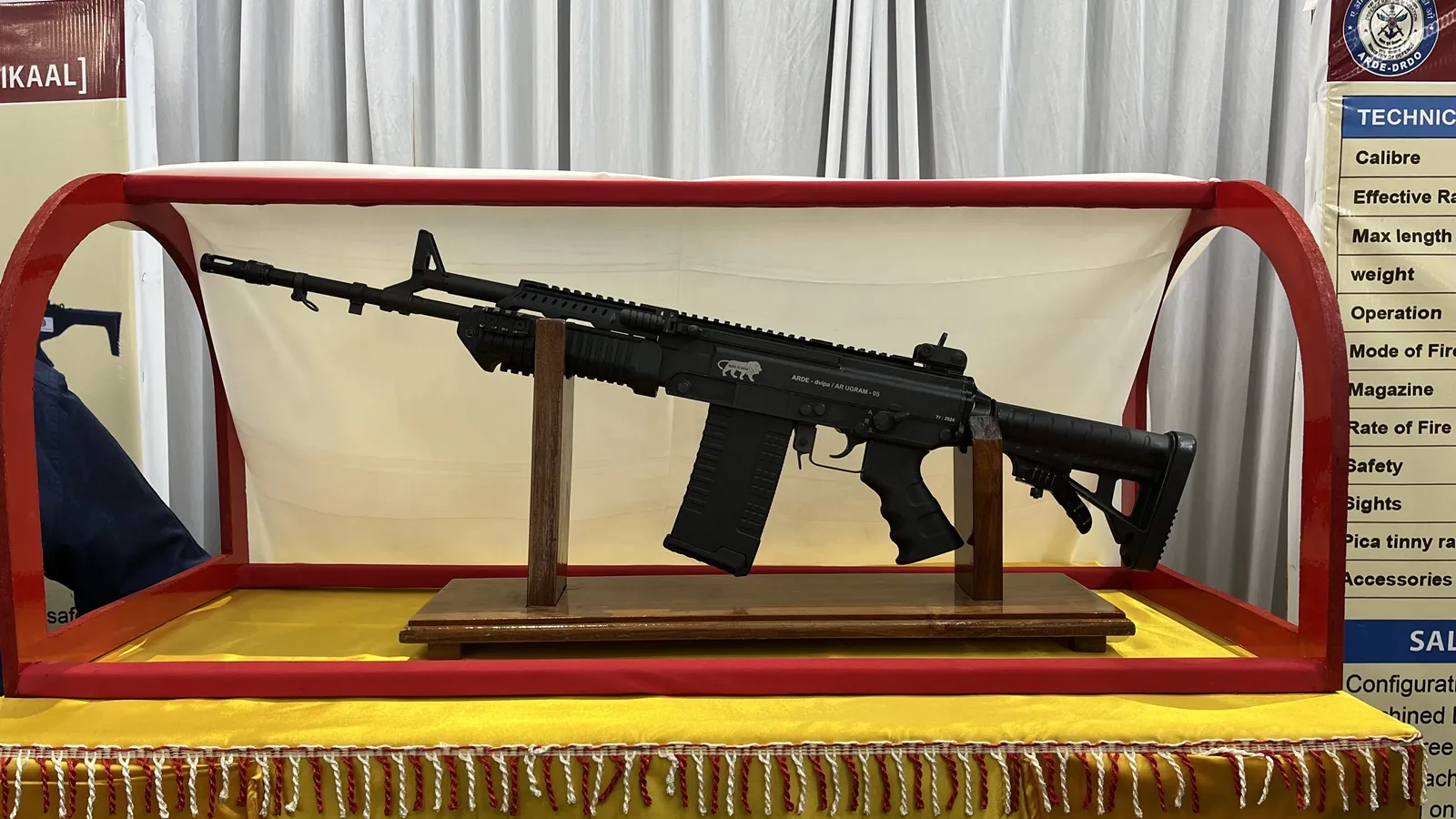The black UGRAM 7.62 mm battle rifle on a wooden display stand inside a red showcase, with an ARDE-DRDO specification board beside it, at its unveiling in Pune.