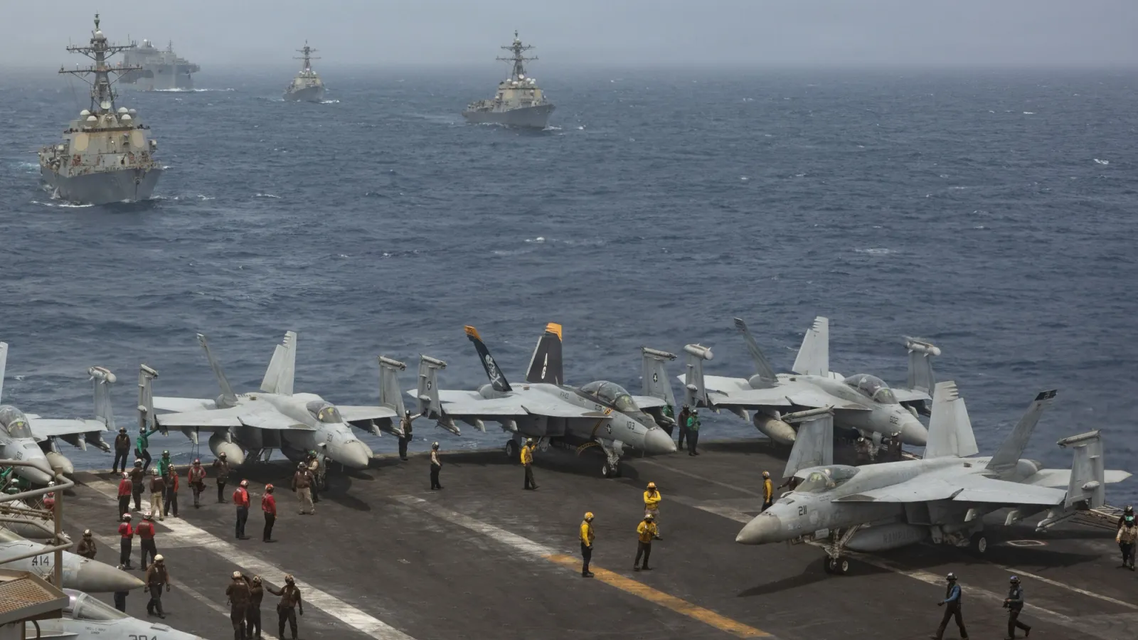 F/A-18 Super Hornets and flight-deck crew on a US Navy aircraft carrier in the Arabian Sea, with guided-missile destroyers and an amphibious warship sailing in formation behind it, 30 June 2026.