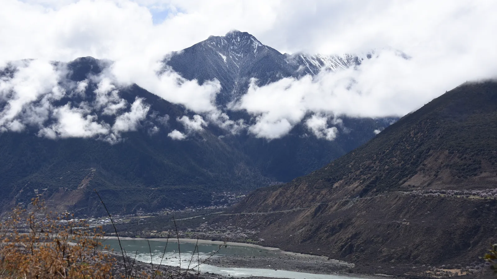 A snow-capped Himalayan peak rising above the Yarlung Tsangpo Grand Canyon in Tibet, with the river winding through the gorge below.